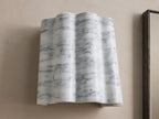 A wall-mounted sconce with a wavy, white marble finish featuring gray veining, installed on a beige wall beside a rectangular mirror.