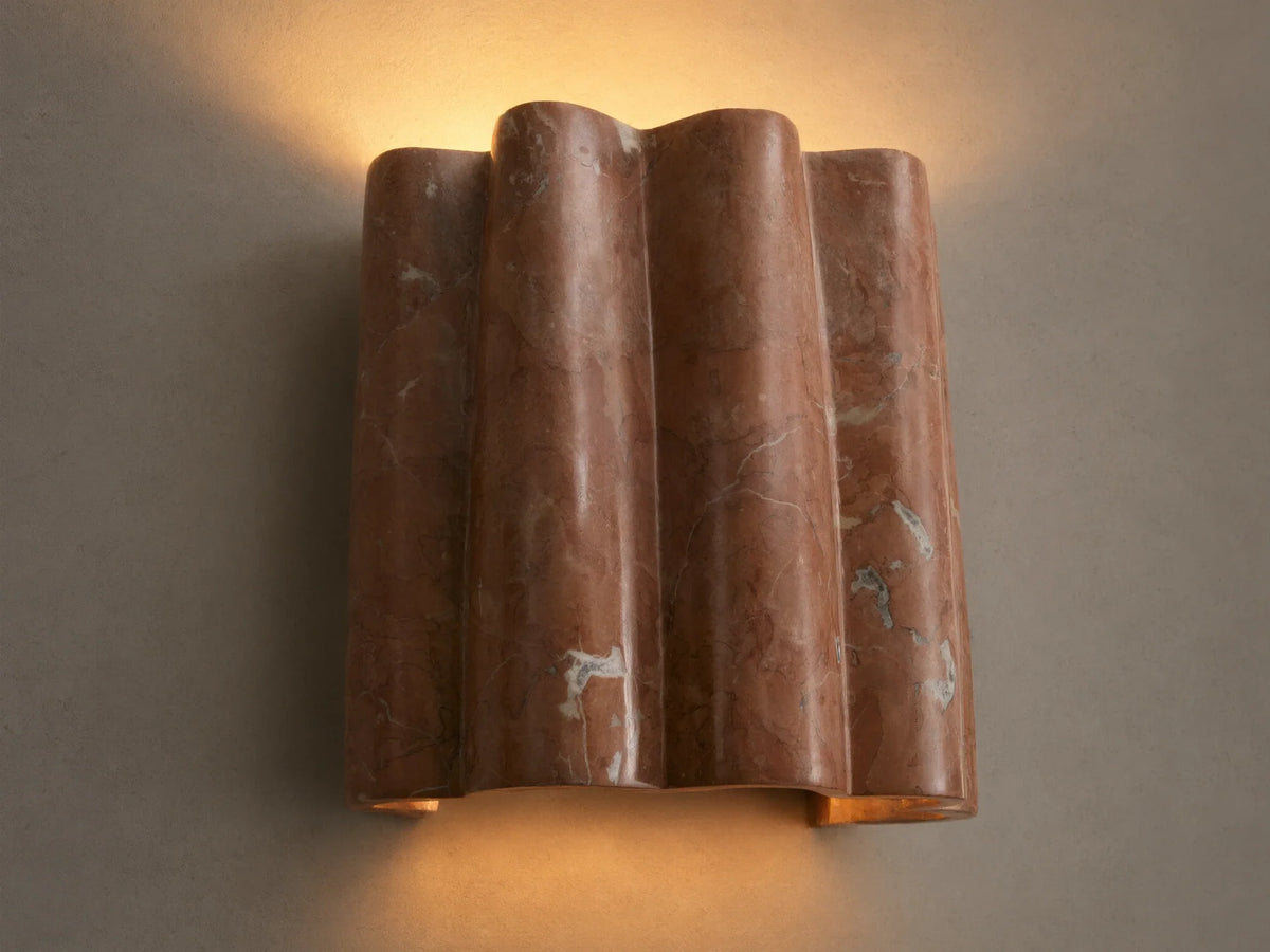 Faine Sconce, a wavy reddish-pink marble wall light with natural white veining, casts a warm glow upwards and downwards.