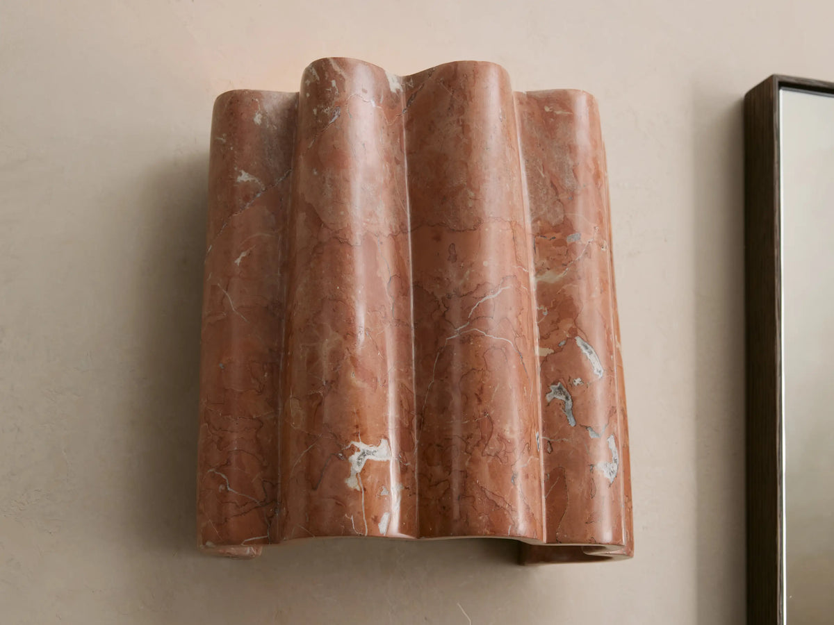 Faine Sconce, a wavy reddish-brown marble wall light with white and grey veins, mounted on a light beige wall.