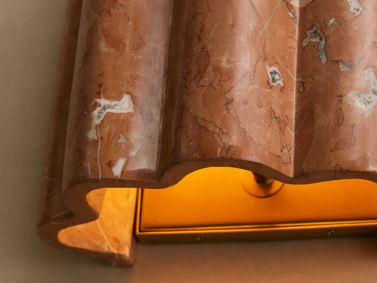Faine Sconce, close-up of a fluted pinkish-red marble sconce with white veins, glowing warm yellow light from its brass base.