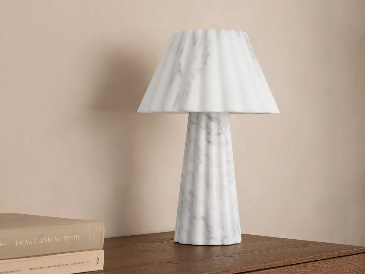 A marble table lamp with a pleated design emits soft light, placed on a wooden surface beside two stacked books.