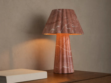 A pink marble table lamp with a fluted base and conical shade casts a warm light on a wooden surface next to a closed book.
