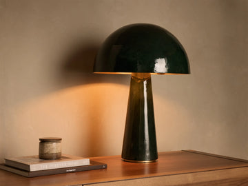 Dark green mushroom-shaped table lamp with brass interior shade, on a wooden surface, emitting warm light.