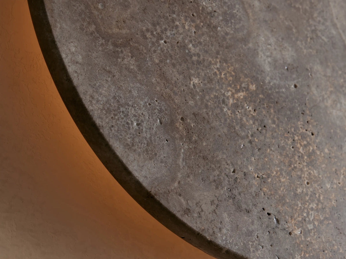Odette Outdoor Sconce. Circular natural stone sconce, mottled grey with brown and lighter gold tones, on a beige wall.