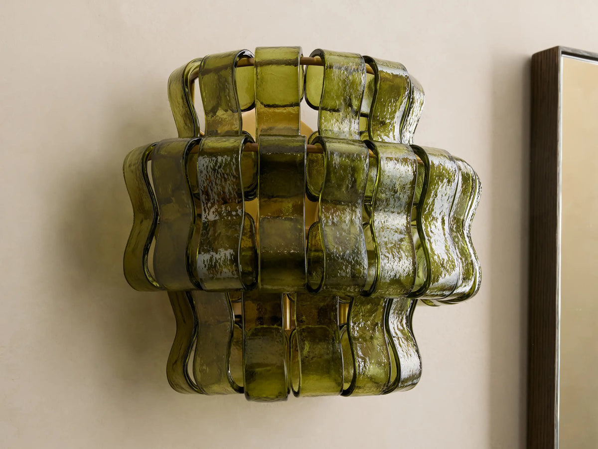 Ghiaccio Sconce, a sculptural wall light with wavy, layered dark green curved glass panels, illuminated with a warm yellow glow on a light wall.