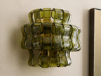 Ghiaccio Sconce, a sculptural wall light with wavy, layered dark green curved glass panels, illuminated with a warm yellow glow on a light wall.
