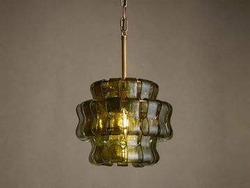 Ghiaccio Pendant, a tiered dark green wavy curved glass pendant light with brass chain, glowing warmly against a neutral wall.