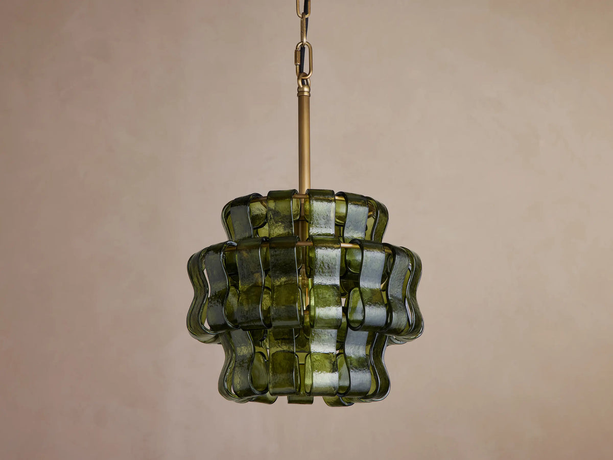 Ghiaccio Pendant, a tiered dark green wavy curved glass pendant light with brass chain, glowing warmly against a neutral wall.