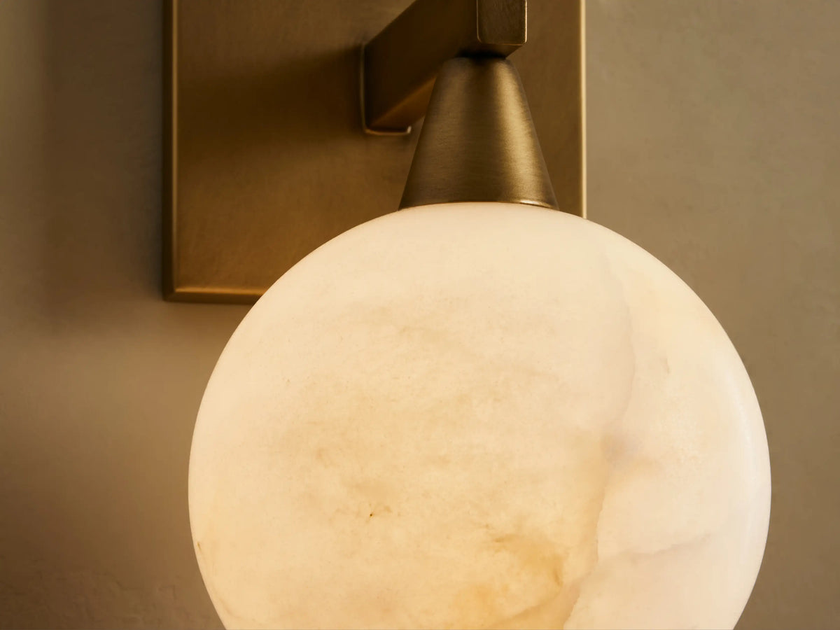 Front view of a brass wall sconce with a square backplate and round milky white glass shade, on a light wall next to a mirror.