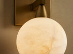 Front view of a brass wall sconce with a square backplate and round milky white glass shade, on a light wall next to a mirror.