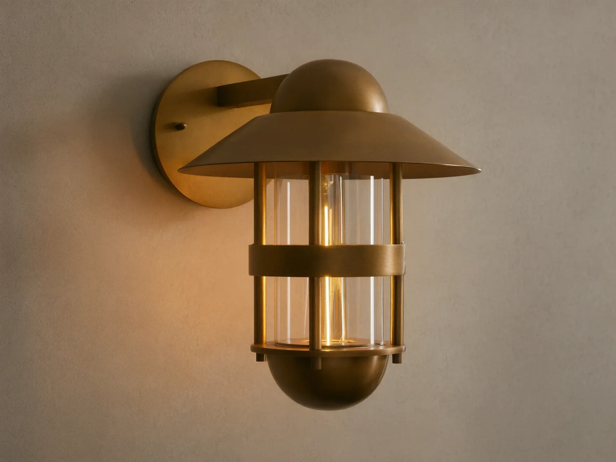 Illuminated Hyannis Outdoor Sconce, antique brass, nautical style with glass housing and visible bulb, on a light wall.
