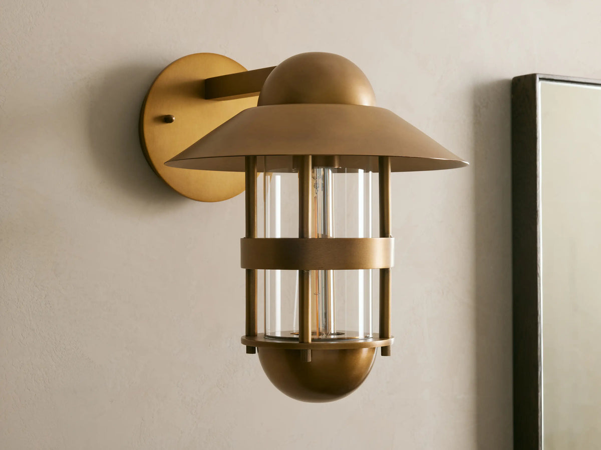 Illuminated Hyannis Outdoor Sconce, antique brass, nautical style with glass housing and visible bulb, on a light wall.