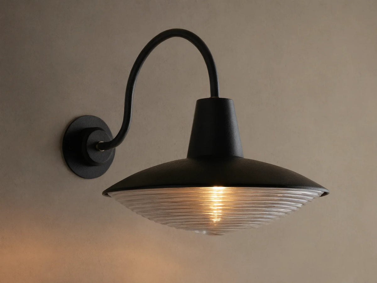 Close-up of Bennett Outdoor Sconce, black wall-mounted with a curved arm. Ribbed clear glass diffuser glows with warm light.