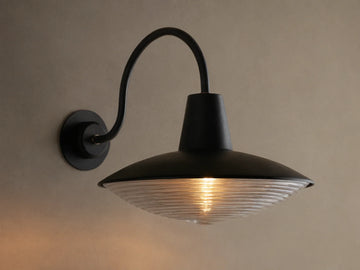 Close-up of Bennett Outdoor Sconce, black wall-mounted with a curved arm. Ribbed clear glass diffuser glows with warm light.