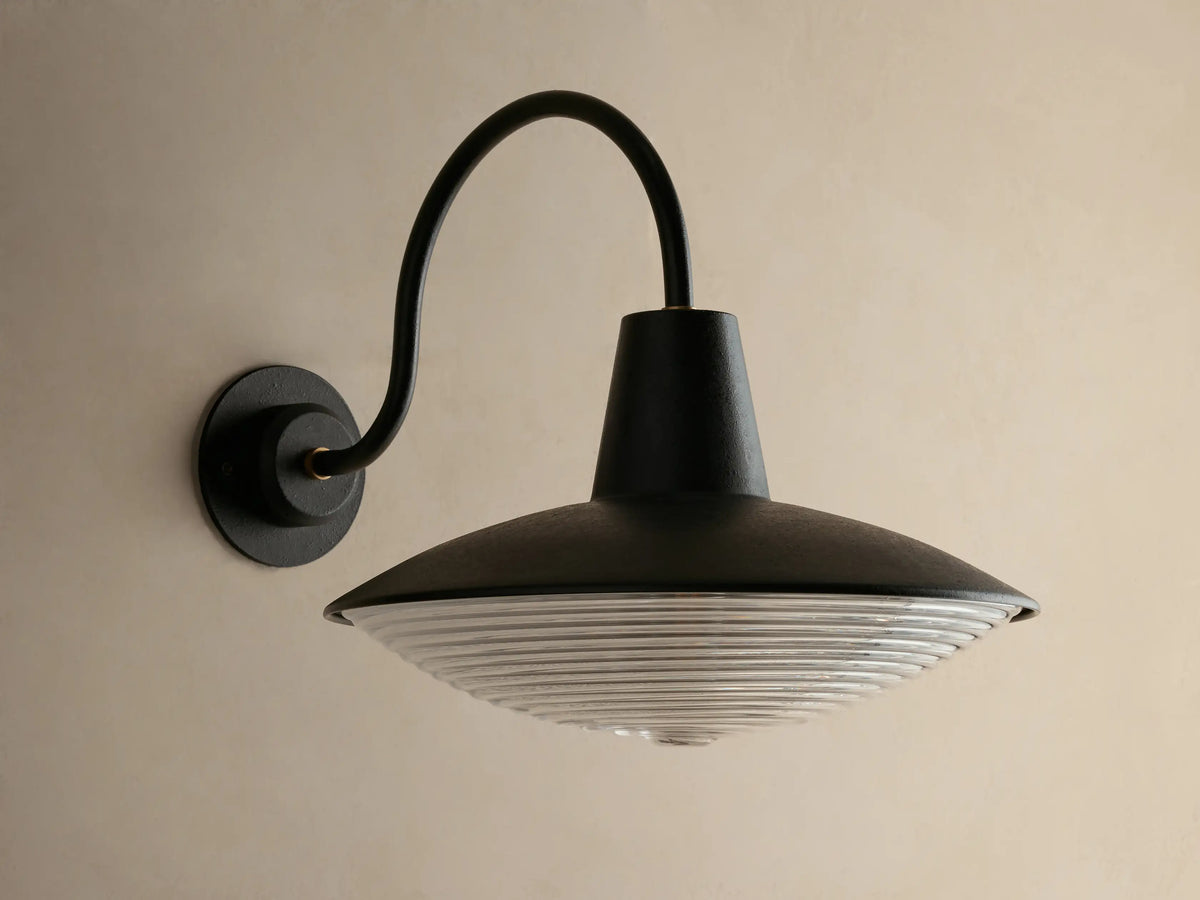 Close-up of Bennett Outdoor Sconce, black wall-mounted with a curved arm. Ribbed clear glass diffuser glows with warm light.