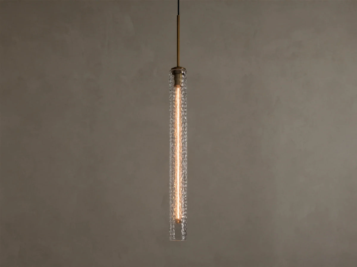 Vertical view of a lit pendant light with a clear textured glass cylinder, a warm filament bulb, and a brass-colored top fixture.