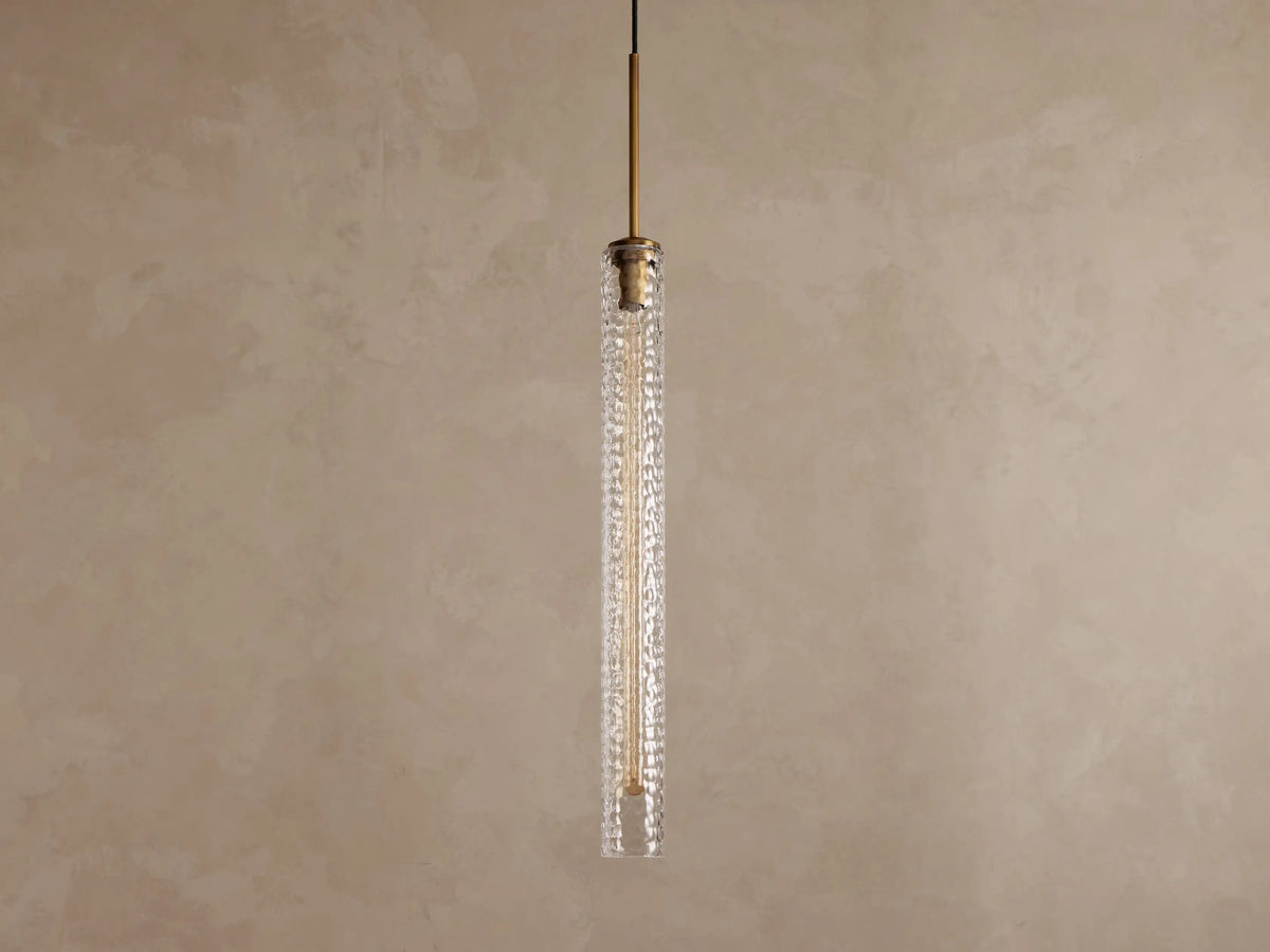 Vertical view of a lit pendant light with a clear textured glass cylinder, a warm filament bulb, and a brass-colored top fixture.