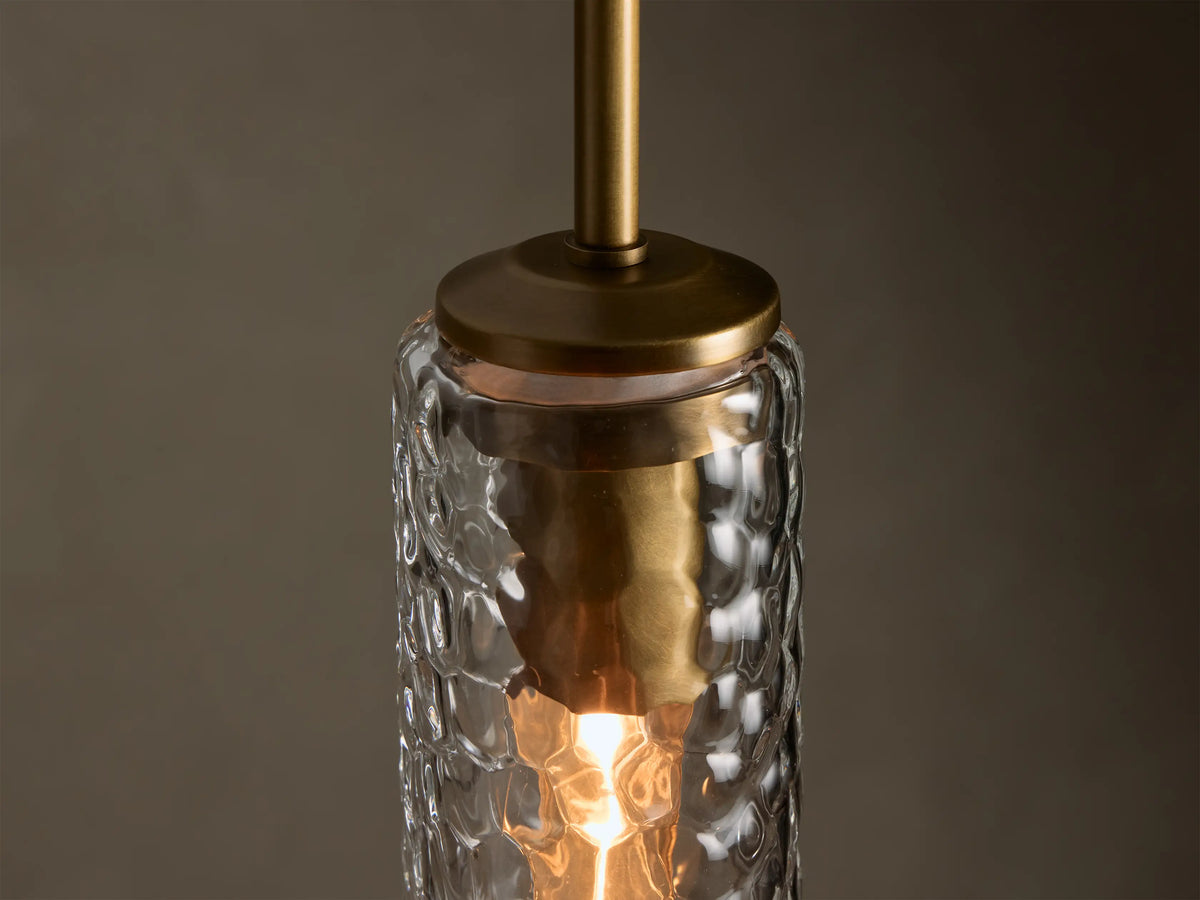 Clear glass pendant light, featuring a vertical hammered cylindrical shade, a brass top, and a warm glow.