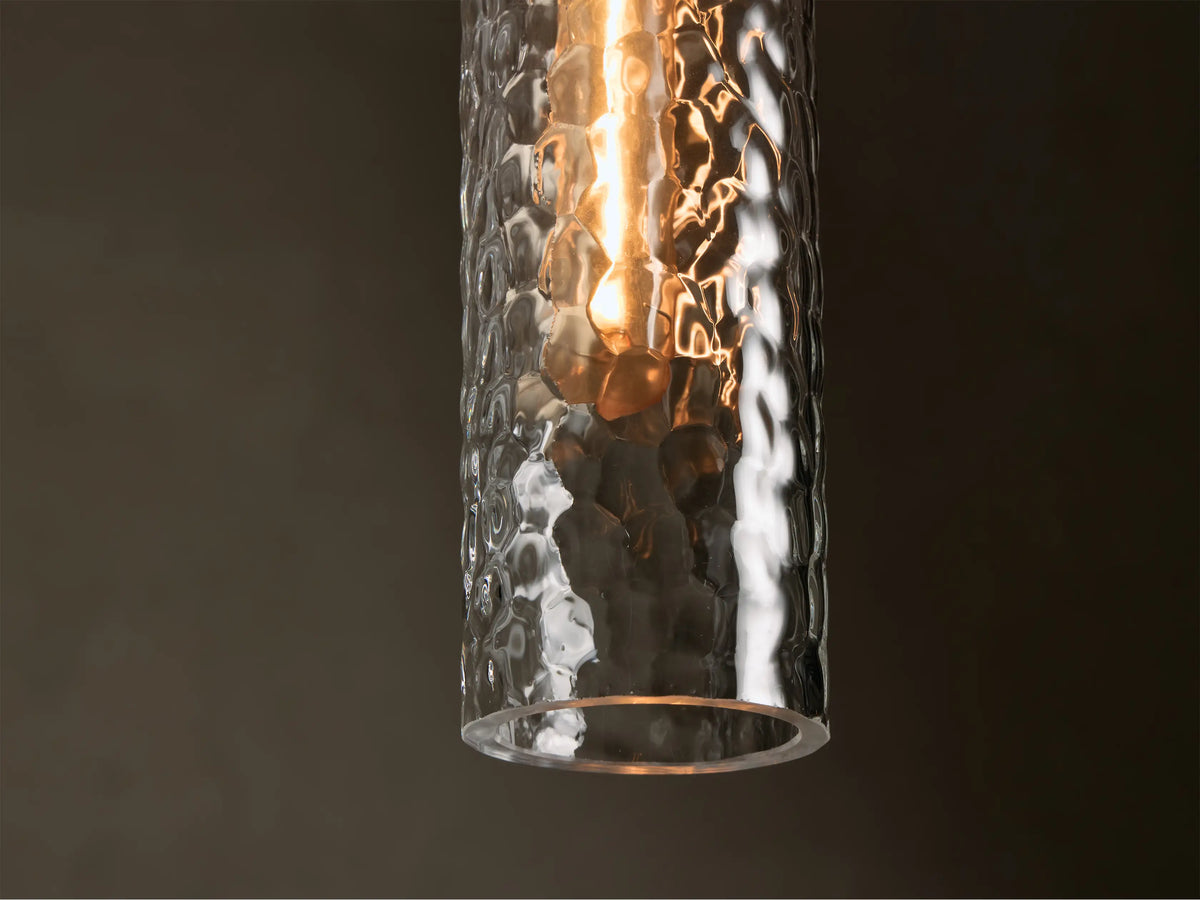 Close-up of a clear hammered glass pendant light with antique brass cap, showing a warm glowing bulb inside.