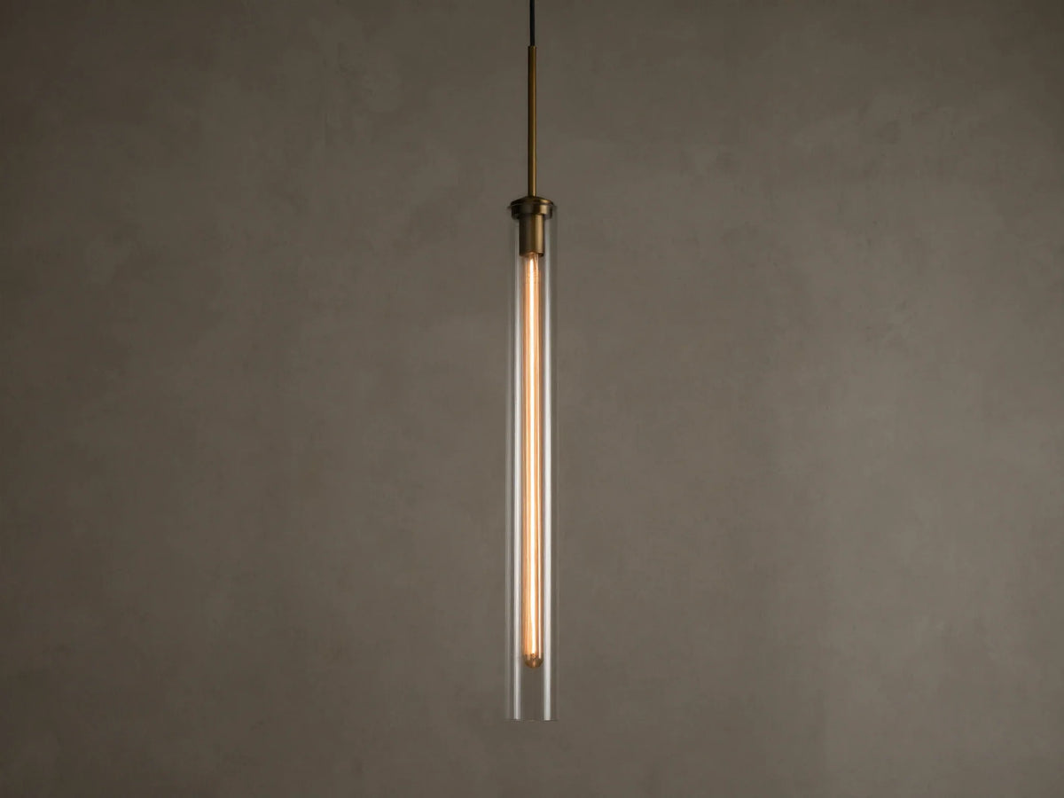 Front view of a modern brass and clear glass pendant light fixture with a long, illuminated filament bulb inside.