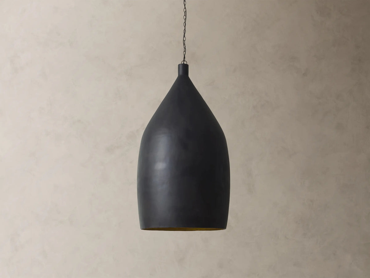 Fria Cone Pendant, a matte black conical pendant light with a golden interior glow, suspended by a chain against a textured wall.