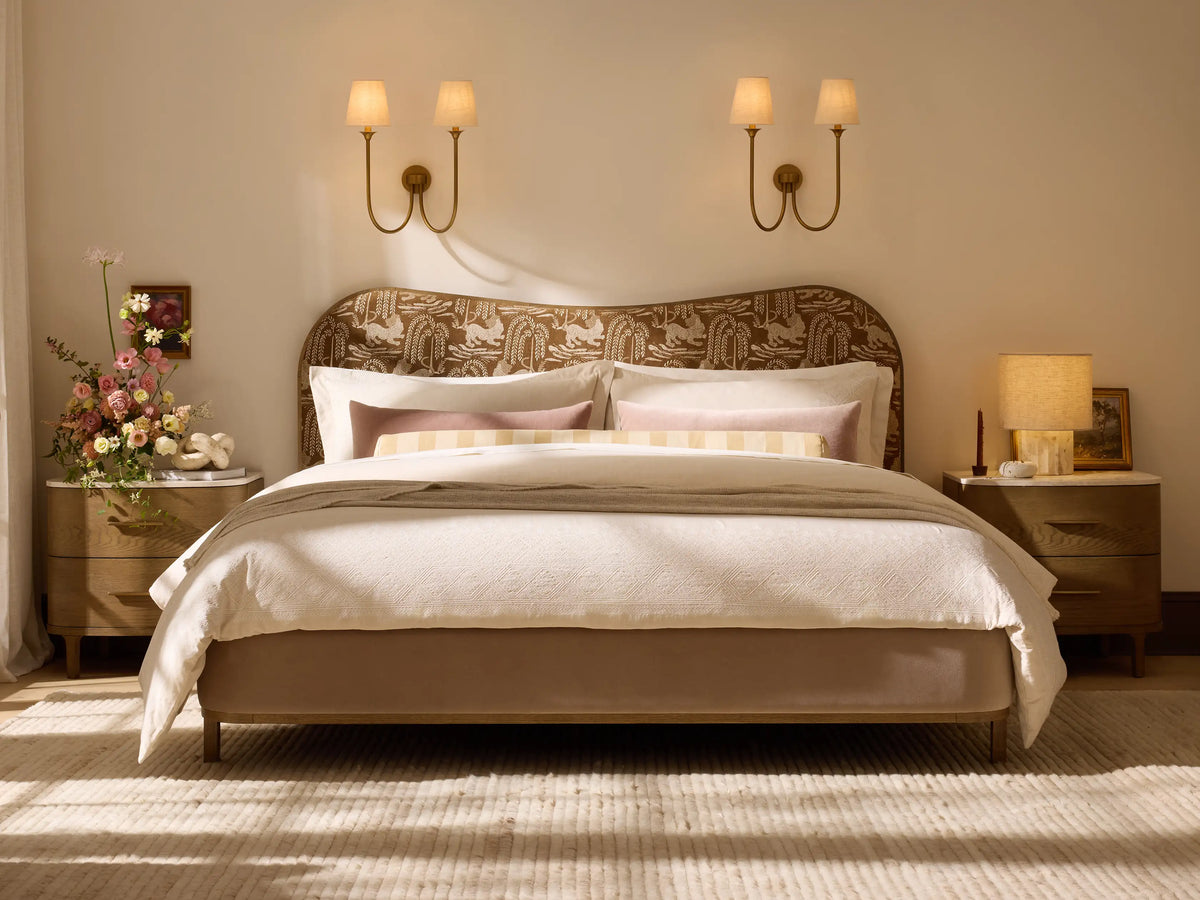 Saige Sconce. Dual wall light with brass finish curved arms, off-white fabric shades glowing warmly on a light wall.