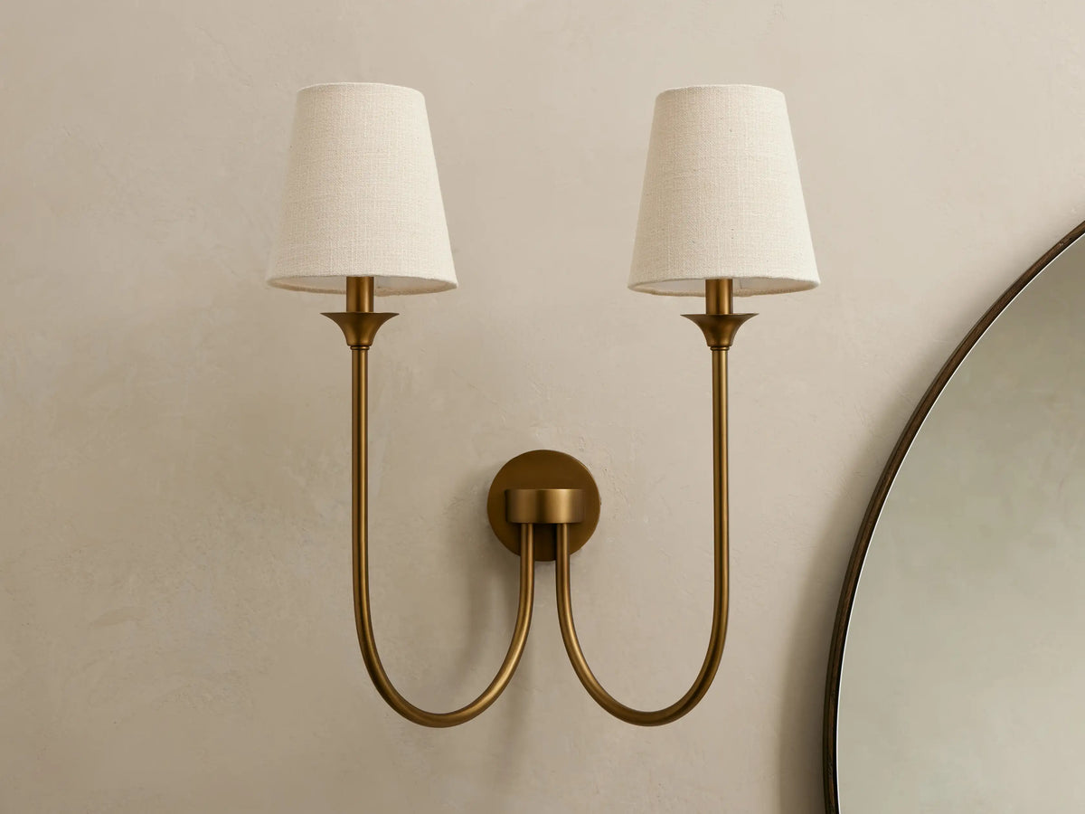Saige Sconce: Dual brass-finish wall sconce with curved arms, two off-white conical fabric shades on a light wall.