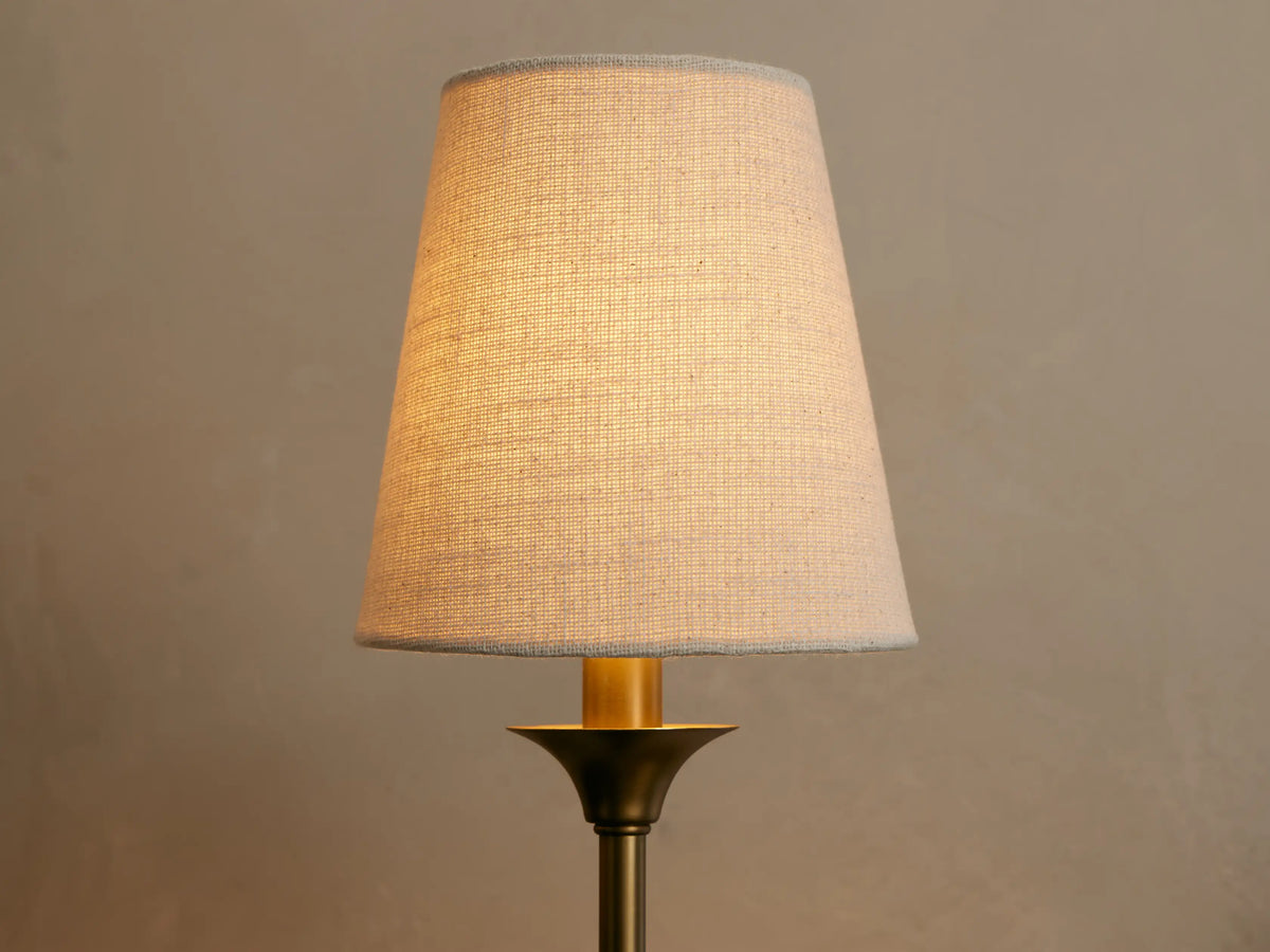 Saige Sconce, close-up of its illuminated off-white linen conical shade and dark brass finish metal body against a neutral wall.