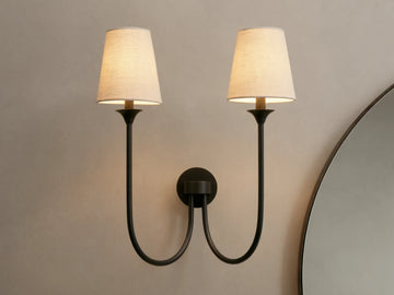 Saige Sconce with two off-white shades & dark metal arms, illuminated on a warm-toned wall next to a mirror.