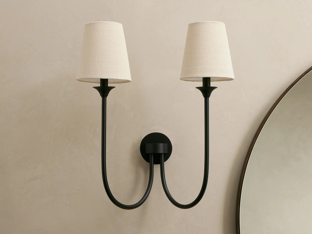 Saige Sconce: Two black metal wall sconces with off-white conical fabric shades on a beige wall, beside a round mirror.