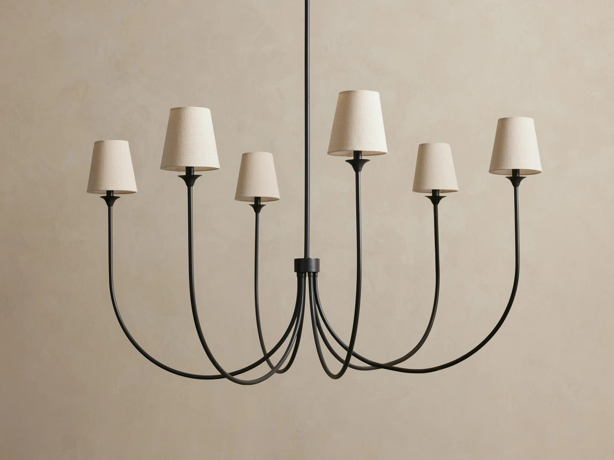 Saige Round Chandelier with 6 curved dark bronze arms & off-white tapered fabric shades, hanging against a light wall.