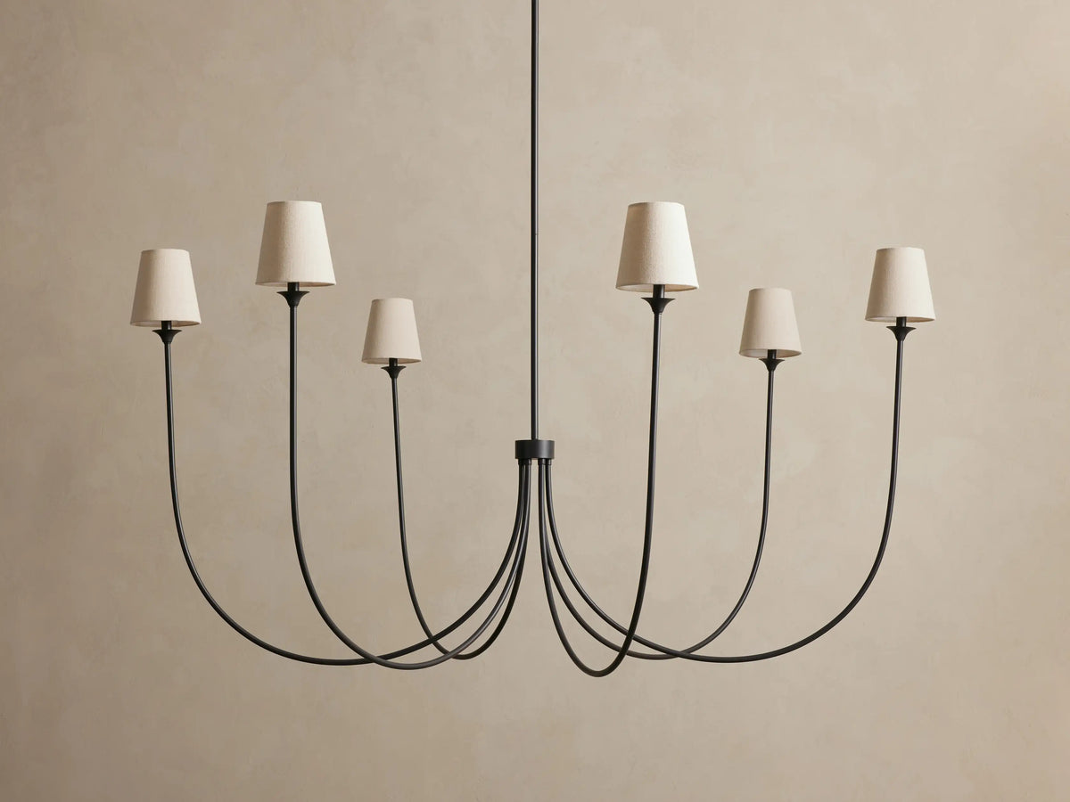 Saige Round Chandelier with 6 dark bronze curved arms, each topped with an off-white fabric shade, against a beige wall.