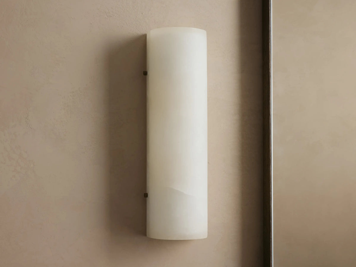 A cylindrical wall sconce light with a soft, warm glow. It has a matte cream finish and is mounted on a textured beige wall.