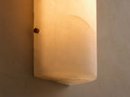 A cylindrical white alabaster wall sconce mounted vertically on a light beige textured wall, adjacent to a mirror.
