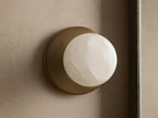 A round, white alabaster wall sconce with a soft glow, set against a textured beige wall. The base is a matte brass finish.