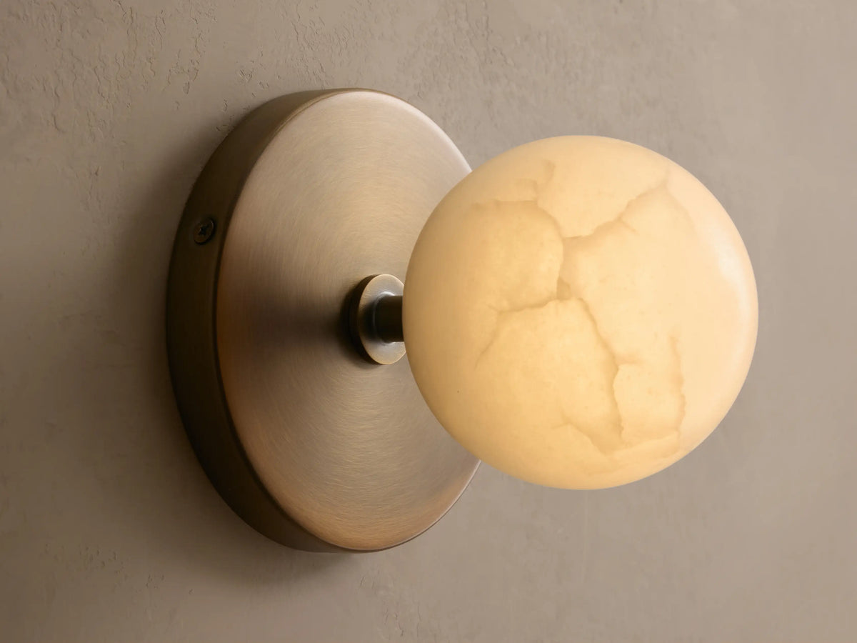 A wall-mounted, spherical light fixture with a warm brass base and a white, frosted glass shade, set against a textured beige wall.