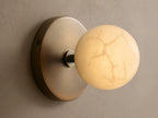 A wall-mounted, spherical light fixture with a warm brass base and a white, frosted glass shade, set against a textured beige wall.