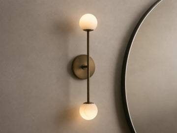 Bronze wall sconce with two frosted glass globes, vertically aligned. Mounted on a light beige wall, near a round mirror.
