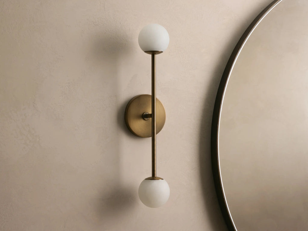 Bronze wall sconce with two frosted glass globes, vertically aligned. Mounted on a light beige wall, near a round mirror.