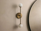 Bronze wall sconce with two frosted glass globes, vertically aligned. Mounted on a light beige wall, near a round mirror.