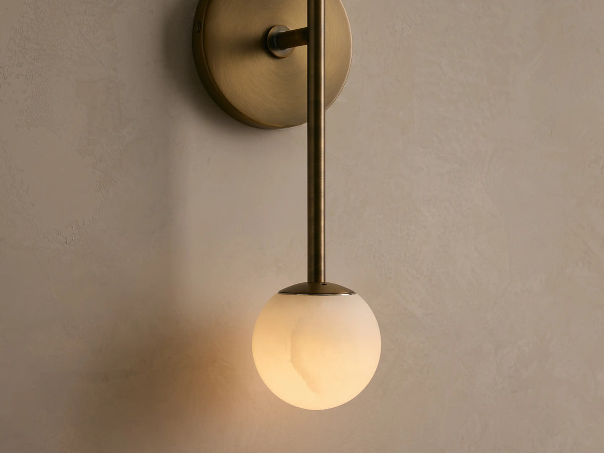 Vertical wall sconce with two spherical white bulbs, featuring a brass base and rod, next to a round mirror on a beige wall.