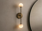 Bronze wall sconce with two frosted glass globes, vertically aligned. Mounted on a light beige wall, near a round mirror.