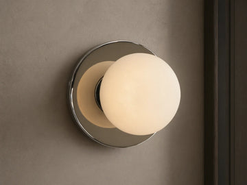 A round wall sconce with a frosted glass globe emits warm light, mounted on a smooth circular metal base, against a beige wall.