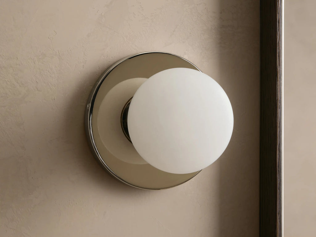 A round wall sconce with a frosted glass globe emits warm light, mounted on a smooth circular metal base, against a beige wall.