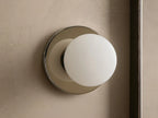 A round wall sconce with a frosted glass globe emits warm light, mounted on a smooth circular metal base, against a beige wall.