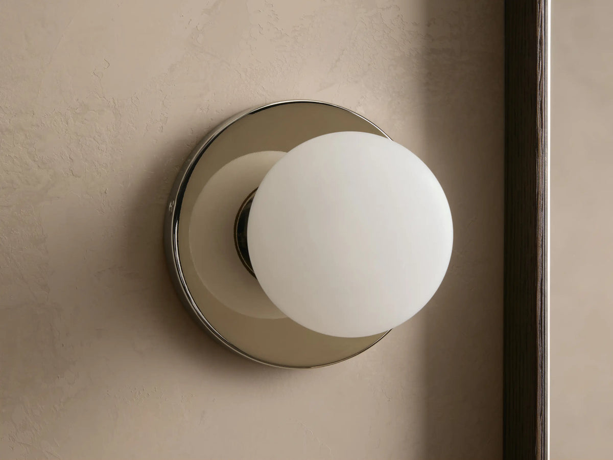 A round wall sconce with a frosted glass globe emits warm light, mounted on a smooth circular metal base, against a beige wall.