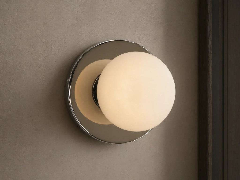 A round wall sconce with a frosted glass globe emits warm light, mounted on a smooth circular metal base, against a beige wall.