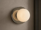 A round wall sconce with a frosted glass globe emits warm light, mounted on a smooth circular metal base, against a beige wall.