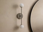 Wall-mounted lamp with a sleek metal fixture, featuring two spherical frosted glass bulbs, vertically aligned against a neutral wall.