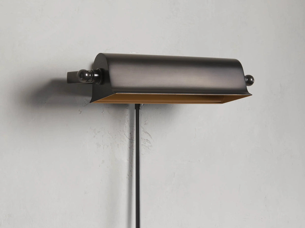 Tremont Bronze Library Sconce with a curved, metallic bronze shade, mounted on a wall, with a thin vertical pole.